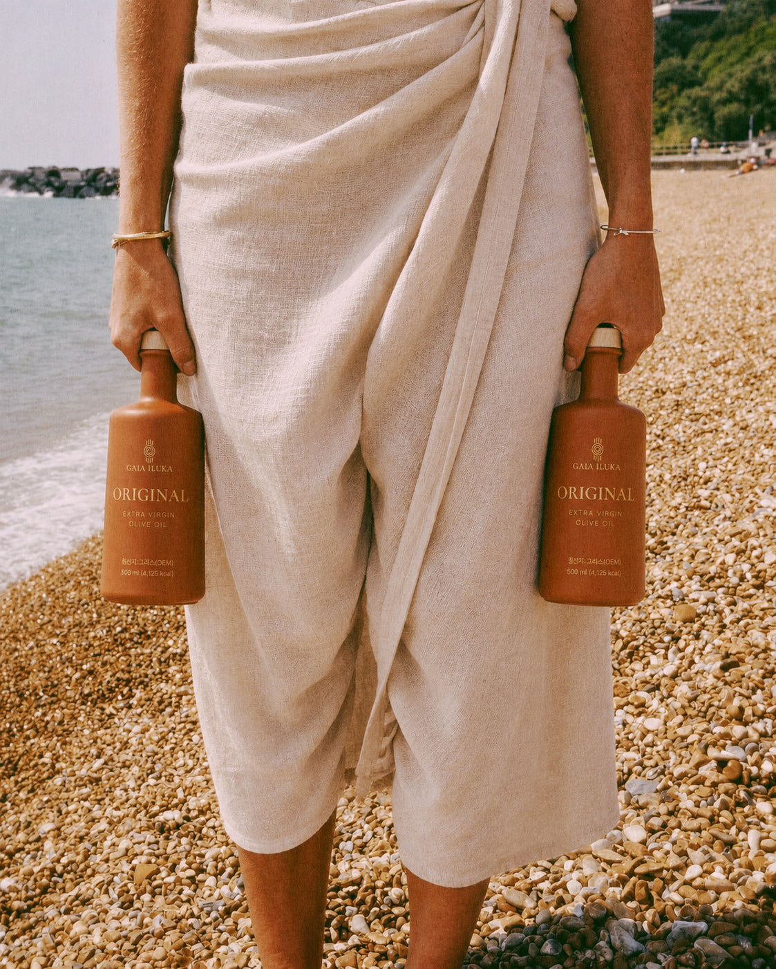 A woman in linen carrying two Gaia Iluka Original bottles on a pebble beach
