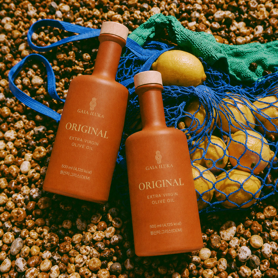 Two Gaia Iluka Original bottles on beach pebbles beside a net bag of lemons