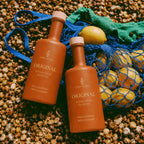 Two Gaia Iluka Original bottles on beach pebbles beside a net bag of lemons
